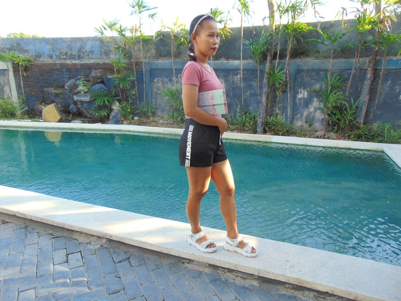 Pu&ograve; includere: Una donna indossa un top corto a fantasia rosa e bianca, pantaloncini neri con la scritta "MOVEMENT" in bianco e sandali bianchi. &Egrave; in piedi vicino a una piscina.