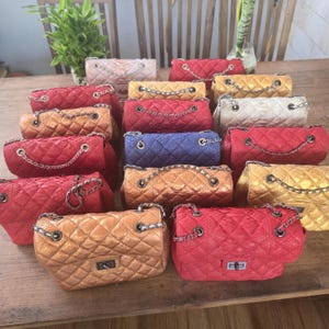 Könnte beinhalten: Eine Sammlung rechteckiger Umhängetaschen mit gesteppter Textur und metallischem Finish in verschiedenen Farben wie Rot, Gold, Blau und Weiß. Jede Tasche verfügt über einen silberfarbenen Kettenriemen und einen rechteckigen Drehverschluss.