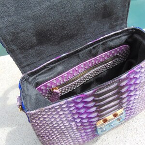 May include: Purple snakeskin handbag with a black lining and a gold zipper. The bag has a flap closure with a gold clasp.
