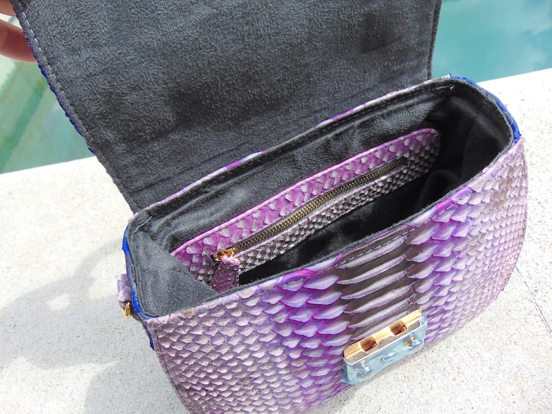May include: Purple snakeskin handbag with a black lining and a gold zipper. The bag has a flap closure with a gold clasp.