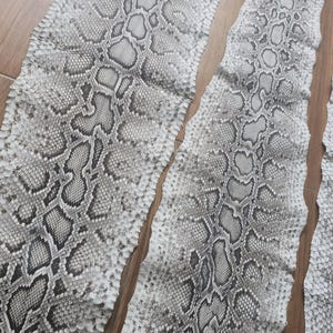 以下が含まれることがあります： グレーと白の色合いの自然な模様が施された、ヘビ革の複数のピース。革は木製の表面に配置されており、詳細な鱗のパターンと質感を際立たせています。