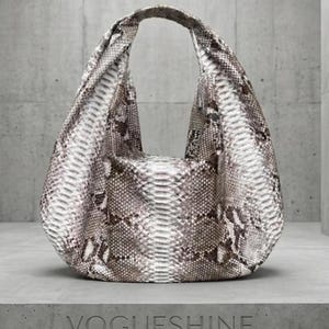 Puede incluir: Un bolso de estilo hobo con un patrón de piel de serpiente texturizado en tonos gris, blanco y marrón. El bolso tiene un asa integrada única y una silueta redondeada y holgada. La palabra VOGUESHINE está grabada en la base debajo del bolso.