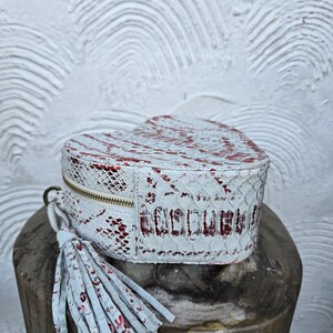 Pu&ograve; includere: Una borsa a forma di cuore con un motivo a pelle di serpente bianco e rosso. La borsa ha una cerniera dorata e un nappa bianca. La borsa &egrave; appoggiata su una superficie di legno.