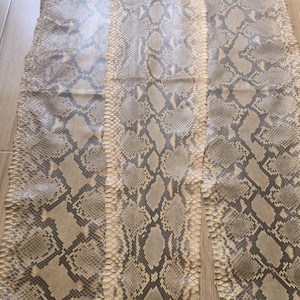 Könnte beinhalten: Drei Pythonhaut-Häute mit einem natürlichen Muster aus grauen, braunen und cremefarbenen Schuppen. Die Häute liegen auf einer Holzoberfläche und zeigen die Textur- und Farbvariationen.