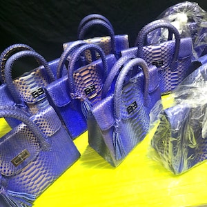 May include: A collection of structured handbags featuring a vibrant blue and metallic gold snakeskin-like texture. Each bag includes a top handle, a rectangular flap with a silver-toned metal turn-lock closure, and a decorative tassel detail.