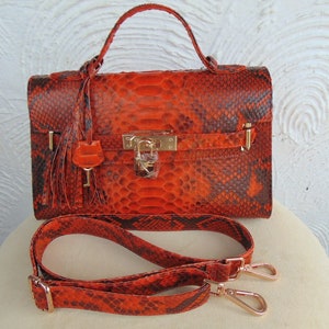 K&ouml;nnte beinhalten: Orangefarbene Schlangenleder-Handtasche mit goldenem Schloss und Schl&uuml;ssel, einem Quastenanh&auml;nger und einem abnehmbaren Schulterriemen.