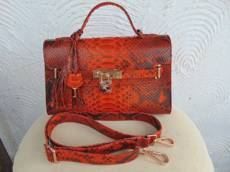 K&ouml;nnte beinhalten: Orangefarbene Schlangenleder-Handtasche mit goldenem Schloss und Schl&uuml;ssel, einem Quastenanh&auml;nger und einem abnehmbaren Schulterriemen.