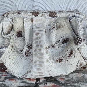 Może przedstawiać: Biała kopert&oacute;wka ze sk&oacute;ry węża z brązowymi oznaczeniami. Torebka wykonana jest z prawdziwej sk&oacute;ry węża i ma unikalny, fakturowany wz&oacute;r.