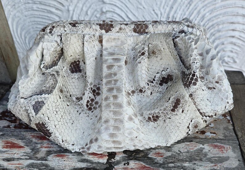 Może przedstawiać: Biała kopert&oacute;wka ze sk&oacute;ry węża z brązowymi oznaczeniami. Torebka wykonana jest z prawdziwej sk&oacute;ry węża i ma unikalny, fakturowany wz&oacute;r.