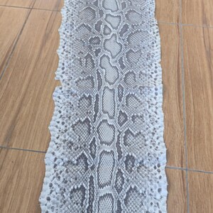 Peut inclure: Une peau de serpent tann&eacute;e, longue et plate, pr&eacute;sentant un motif r&eacute;p&eacute;titif d'&eacute;cailles en forme de losange gris et blanc. La peau est allong&eacute;e et &eacute;troite, affichant une texture naturelle avec un motif central distinct et des bords irr&eacute;guliers.