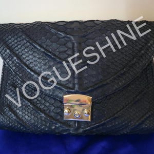 Op de afbeelding: Een donkerblauwe clutch met een textuur, slangachtig patroon. De tas heeft een klepsluiting met een goudkleurige sluiting. De tekst "VOGUESHINE" is over de voorkant van de tas gedrukt.