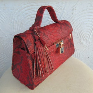 K&ouml;nnte beinhalten: Eine rote Schlangenleder-Handtasche mit einem goldenen Schloss und einem Quastenanh&auml;nger. Die Tasche hat einen oberen Henkel und einen Klappenverschluss.