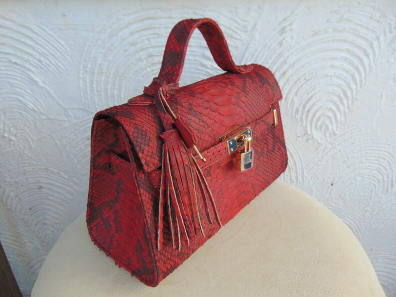 K&ouml;nnte beinhalten: Eine rote Schlangenleder-Handtasche mit einem goldenen Schloss und einem Quastenanh&auml;nger. Die Tasche hat einen oberen Henkel und einen Klappenverschluss.