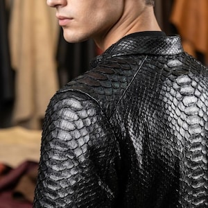 May include: A black jacket featuring a textured, scale-like pattern across the entire surface. The garment has a structured collar and long sleeves, with a glossy finish that highlights the overlapping, reptilian-inspired scale design.