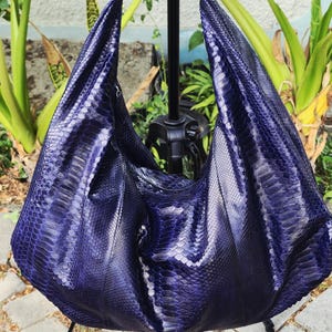 May include: A deep purple hobo bag featuring a textured snakeskin pattern. The bag has a slouchy, crescent-shaped silhouette with a single integrated shoulder strap and a smooth, glossy finish that reflects light across its scales.