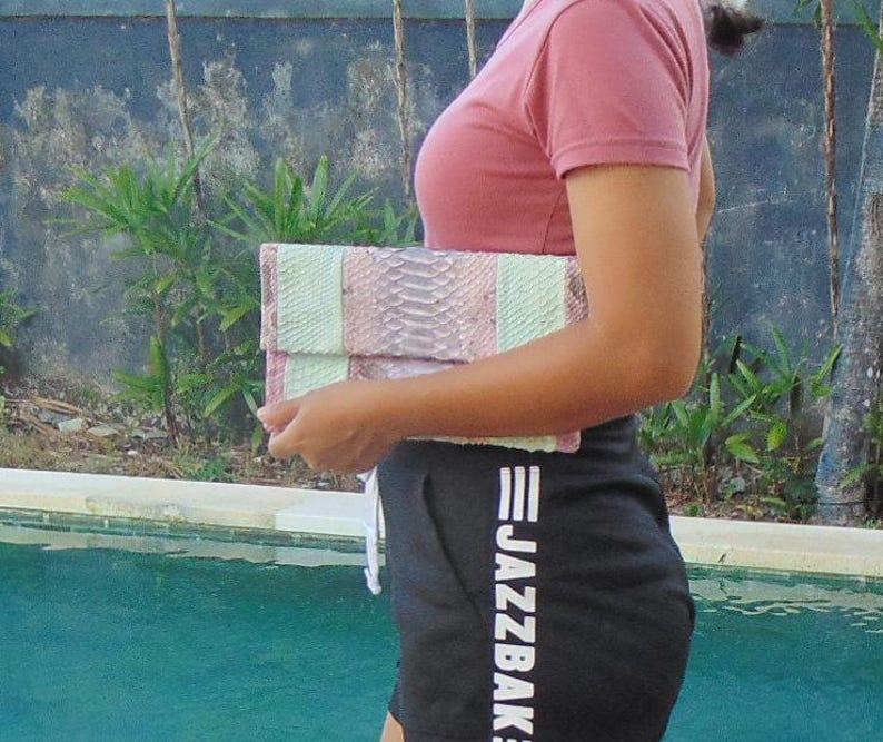 Pu&ograve; includere: Una donna tiene in mano una pochette in pelle di serpente rosa e bianca con un motivo a righe bianche e nere. La pochette ha una chiusura argentata. La donna indossa una camicia rosa e pantaloncini neri con la scritta "JAZZBAK" stampata sul lato.