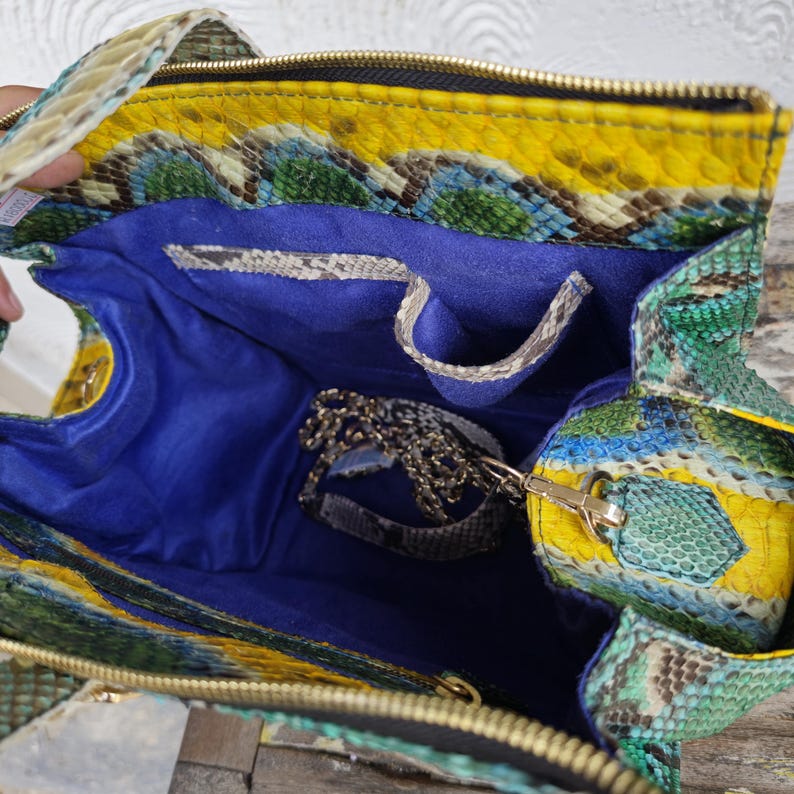 K&ouml;nnte beinhalten: Eine gelbe, gr&uuml;ne und blaue Schlangenleder-Handtasche mit einem blauen Stofffutter und einem goldenen Kettenriemen. Die Tasche ist offen und das Innere ist sichtbar.