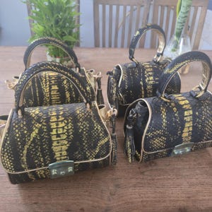 Peut inclure: Trois sacs à main structurés présentant une texture à motif peau de serpent noir et doré. Chaque sac possède une poignée supérieure, une fermeture à fermoir métallique sur le rabat avant et des bordures dorées. Inclut un détail à pompon.