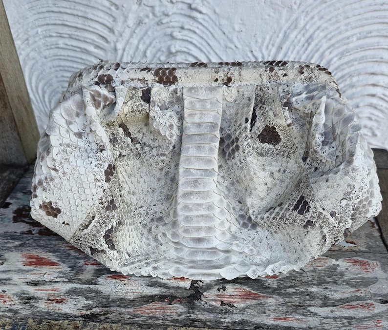 Może przedstawiać: Biała kopert&oacute;wka ze wzorem sk&oacute;ry węża. Torebka ma fakturowaną powierzchnię i lekko zużyty wygląd.