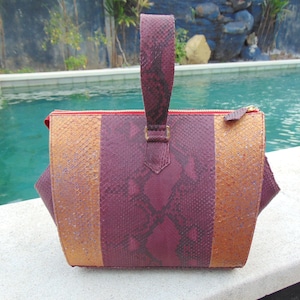 May include: A burgundy and gold snakeskin handbag with a top handle. The bag has a zipper closure and a textured snakeskin pattern.