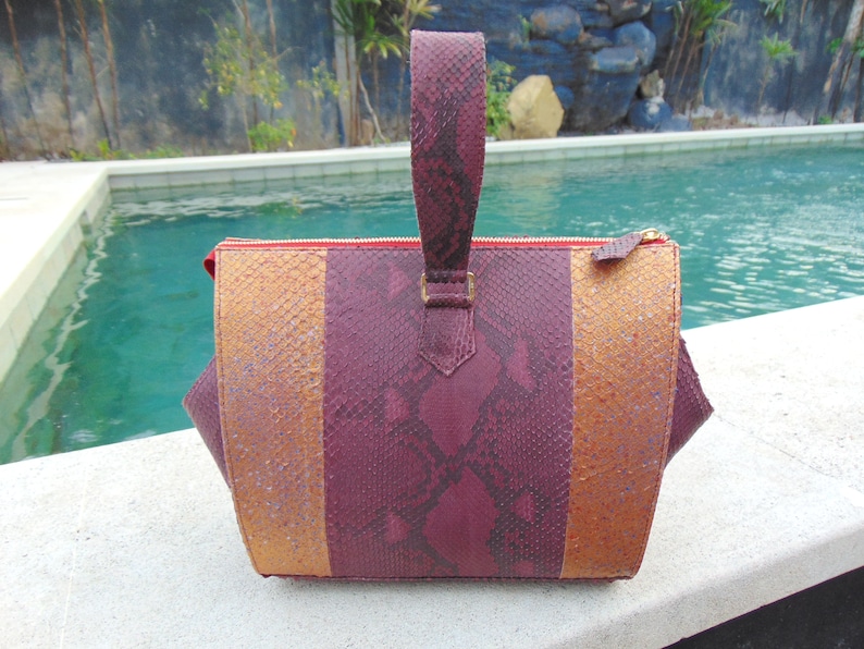 May include: A burgundy and gold snakeskin handbag with a top handle. The bag has a zipper closure and a textured snakeskin pattern.