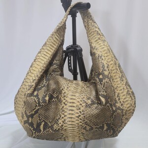 May include: A hobo-style shoulder bag featuring a textured snakeskin pattern in shades of tan, beige, and dark brown. The bag has a wide, integrated shoulder strap and a slouchy, unstructured silhouette with a visible scale texture throughout.