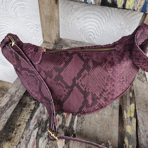 May include: A burgundy fanny pack with a snakeskin pattern. The bag has a curved shape, a zippered top, and a long adjustable strap. Gold-colored hardware accents the bag.