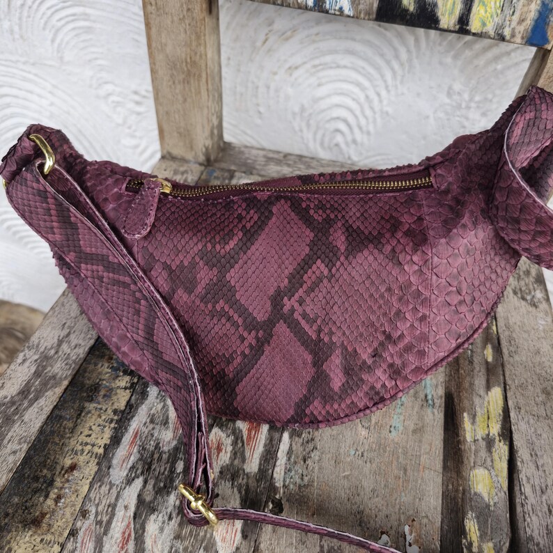 May include: A burgundy fanny pack with a snakeskin pattern. The bag has a curved shape, a zippered top, and a long adjustable strap. Gold-colored hardware accents the bag.
