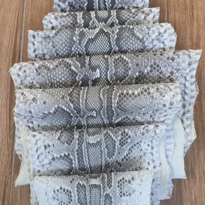 May include: A single piece of natural snakeskin leather featuring a repeating diamond-shaped scale pattern in shades of off-white, light gray, and dark charcoal. The hide is folded in a layered, accordion-style arrangement to display the texture.