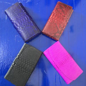 Może przedstawiać: Cztery skórzane portfele typu kopertówka ułożone w wachlarz. Portfele są w odcieniach niebieskiego, czerwonego, czarnego i różowego. Każdy portfel ma fakturę imitującą skórę węża. Portfele leżą na niebieskiej powierzchni.