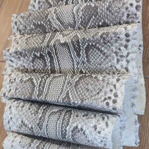 Peut inclure: Une s&eacute;rie de feuilles de cuir de peau de serpent naturel empil&eacute;es et superpos&eacute;es, pr&eacute;sentant un motif distinctif d'&eacute;cailles en forme de losange dans des tons de gris, blanc et noir. Le mat&eacute;riau textur&eacute; est dispos&eacute; en cascade.