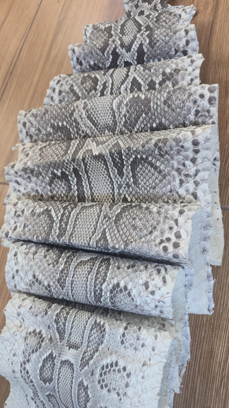 Peut inclure: Une s&eacute;rie de feuilles de cuir de peau de serpent naturel empil&eacute;es et superpos&eacute;es, pr&eacute;sentant un motif distinctif d'&eacute;cailles en forme de losange dans des tons de gris, blanc et noir. Le mat&eacute;riau textur&eacute; est dispos&eacute; en cascade.