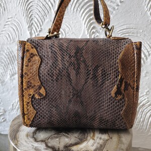 May include: A brown and tan snakeskin handbag with a top handle and a shoulder strap. The bag has a flap closure and a decorative stitch detail.