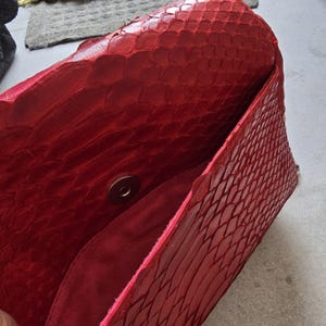 May include: A red clutch bag with a textured, scaly pattern. The interior is a darker red, and the bag has a magnetic snap closure. The bag is open, revealing the inside.