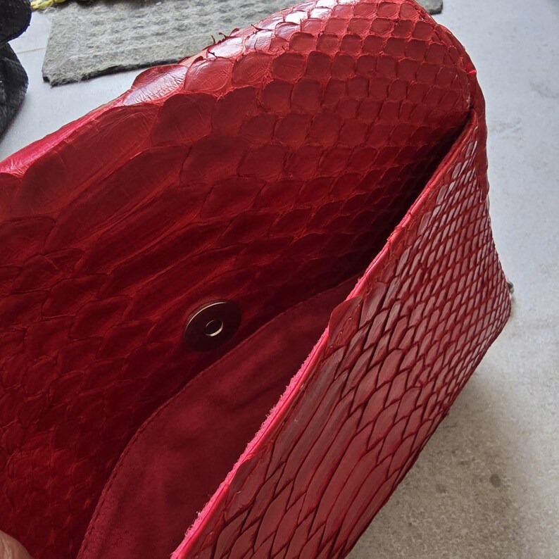 May include: A red clutch bag with a textured, scaly pattern. The interior is a darker red, and the bag has a magnetic snap closure. The bag is open, revealing the inside.
