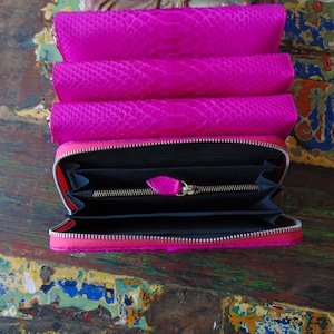 K&ouml;nnte beinhalten: Eine rosa Schlangenleder-Geldb&ouml;rse mit Rei&szlig;verschluss. Die Geldb&ouml;rse ist ge&ouml;ffnet und das Innere ist schwarz.