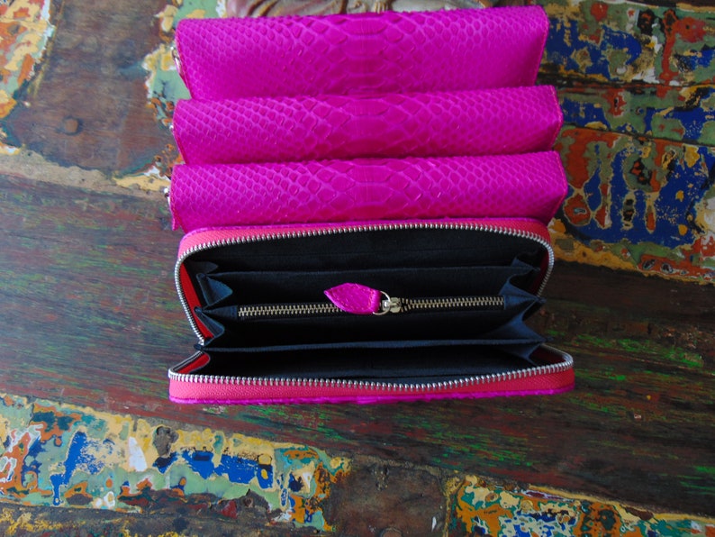 K&ouml;nnte beinhalten: Eine rosa Schlangenleder-Geldb&ouml;rse mit Rei&szlig;verschluss. Die Geldb&ouml;rse ist ge&ouml;ffnet und das Innere ist schwarz.