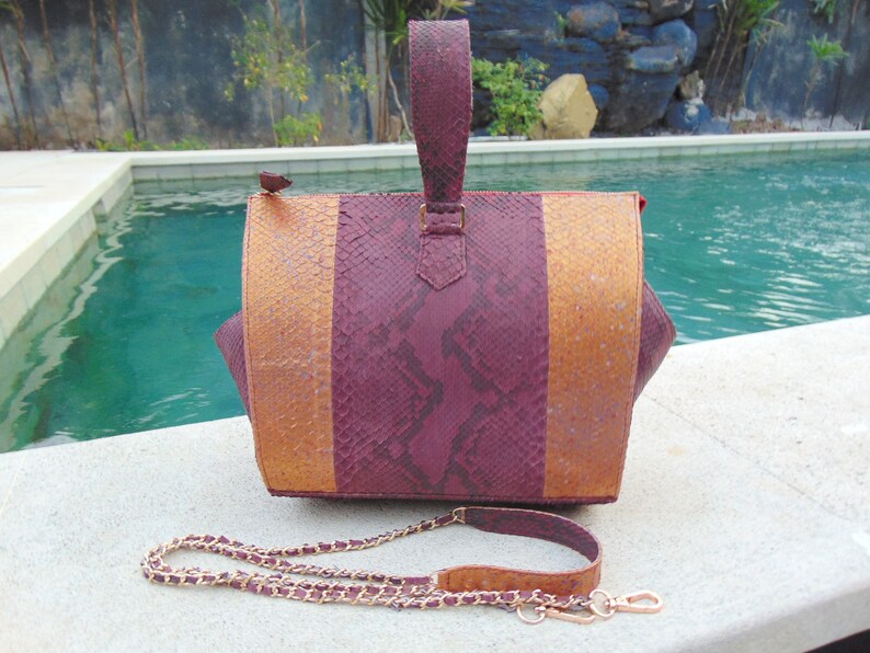 May include: A burgundy and gold snakeskin handbag with a chain strap. The bag has a top handle and is sitting on a concrete surface near a swimming pool.