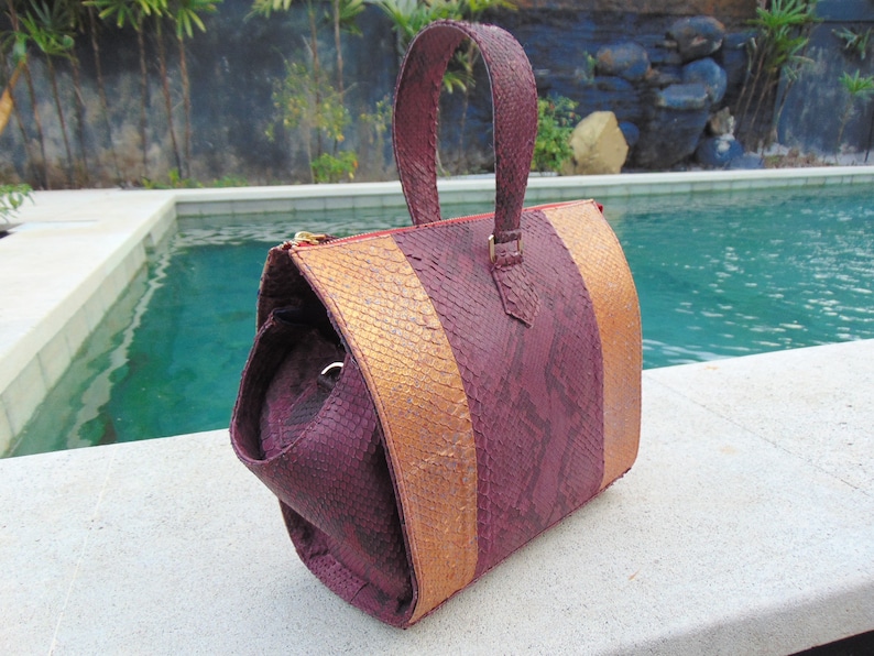 May include: A burgundy and gold snakeskin handbag with a top handle. The bag has a textured snakeskin pattern and a gold zipper.