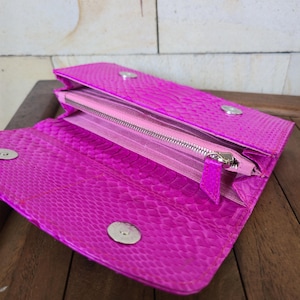 May include: A bright pink wallet with a textured, snake-like pattern. The wallet is open, revealing a pink interior with a zippered compartment and several card slots. Silver-colored snap closures secure the wallet.