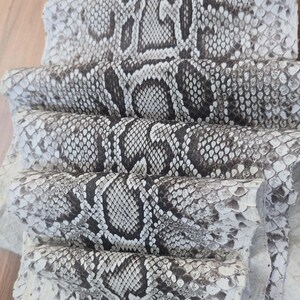 May include: A stack of multiple sheets of genuine snakeskin leather featuring a natural scale pattern in shades of white, grey, and dark brown. The textured material is layered, showcasing the repeating organic diamond-shaped markings.