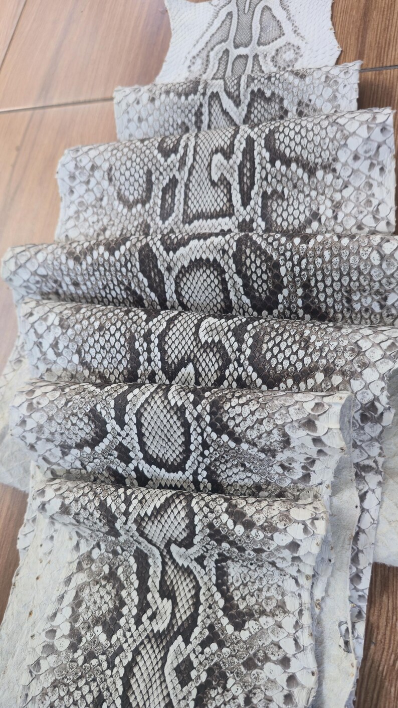 May include: A stack of multiple sheets of genuine snakeskin leather featuring a natural scale pattern in shades of white, grey, and dark brown. The textured material is layered, showcasing the repeating organic diamond-shaped markings.