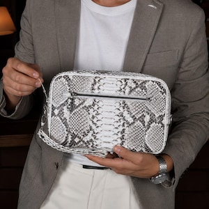 May include: A rectangular white and gray snakeskin clutch with a zipper closure and a wrist strap. The bag is held by a person wearing a gray blazer, white t-shirt, and white pants. A silver watch is visible on the person's wrist.