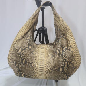 Puede incluir: Un bolso de estilo hobo con un patrón de piel de serpiente texturizada en tonos beige, bronceado y marrón oscuro. El bolso tiene una silueta holgada y desestructurada con una sola correa integrada y una amplia abertura superior.