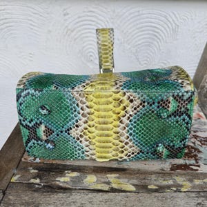 K&ouml;nnte beinhalten: Eine gr&uuml;ne, gelbe und braune Schlangenleder-Handtasche mit einem Henkel oben.