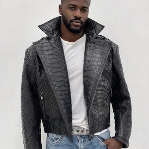 May include: Black leather jacket with a snakeskin pattern, featuring a wide collar and a zipper closure. The jacket is worn over a white t-shirt and blue jeans. A silver belt is visible.