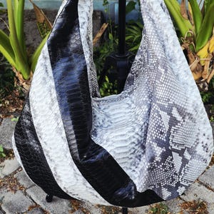 May include: A hobo-style shoulder bag featuring a textured snakeskin pattern in a color palette of black, white, and gray. The bag has a slouchy, unstructured silhouette with a wide integrated shoulder strap and a distinct diagonal color-blocked design.