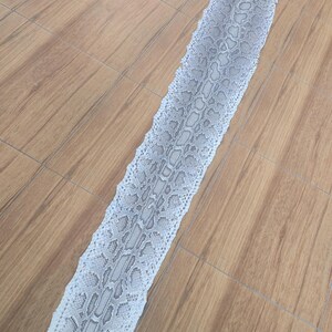 Peut inclure: Une longue mue de serpent pr&eacute;sentant un motif d'&eacute;cailles complexe et r&eacute;p&eacute;titif dans des tons blanc cass&eacute;, gris clair et anthracite. La peau est dispos&eacute;e en une ligne droite et allong&eacute;e, r&eacute;v&eacute;lant une surface naturelle et textur&eacute;e.