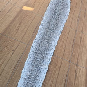 May include: A long, flat, tanned snakeskin featuring a repeating scale pattern in shades of white, light gray, and dark gray. The skin is elongated and narrow, displaying a natural, textured surface with a distinct, symmetrical diamond-like scale arrangement.