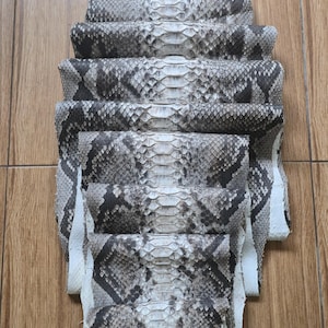 Puede incluir: Pieles de serpiente curtidas apiladas en tonos grises, negros y blancos, con un patrón de escamas natural. Las pieles están dispuestas en una pila vertical, mostrando la textura y el patrón del cuero de reptil. Se muestran sobre una superficie de madera.